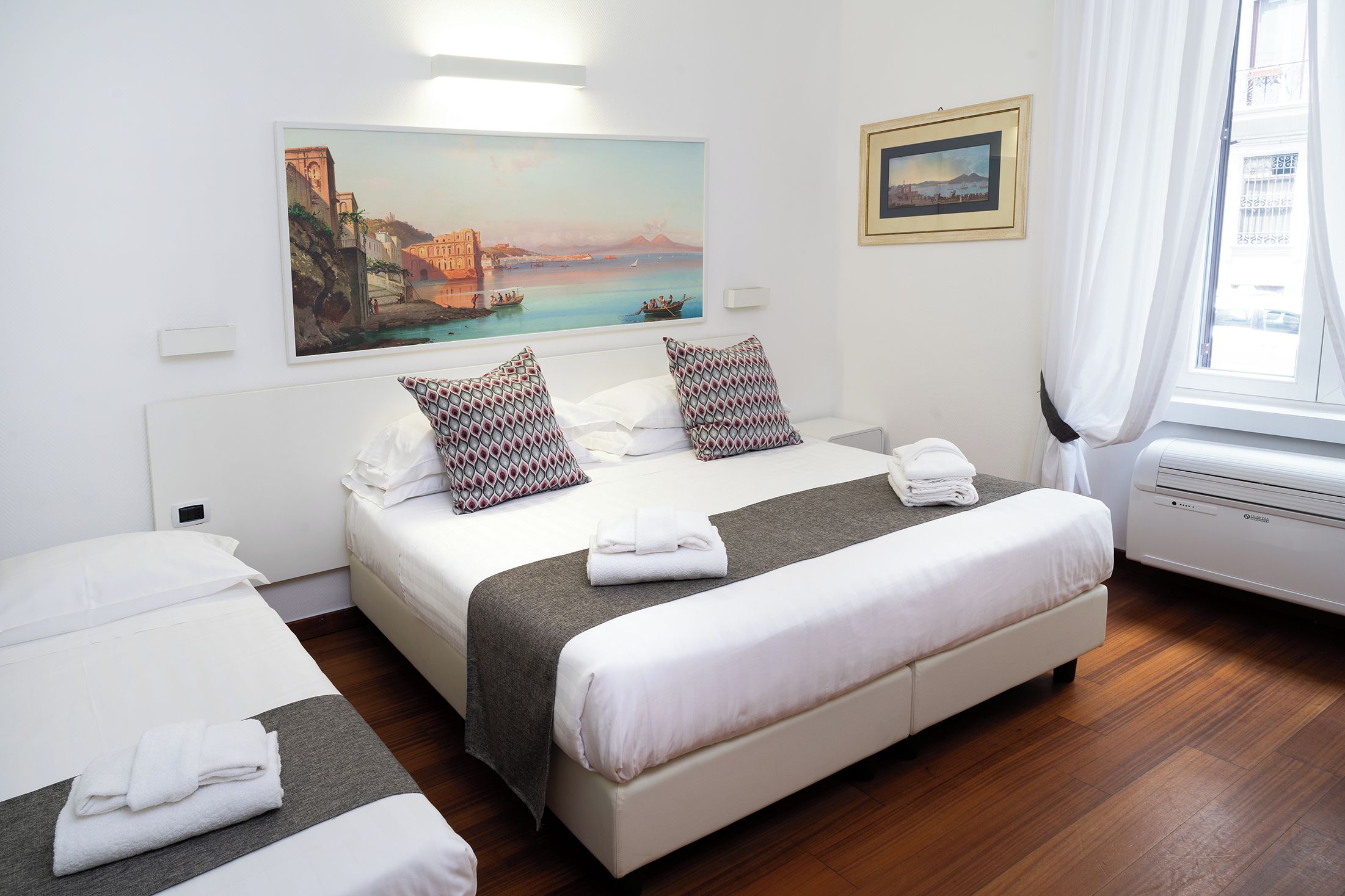 A hotel room with a bed, two pillows, a painting, a window with curtains, and a wall-mounted air conditioner.