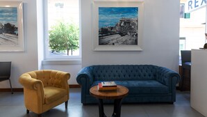 Une pièce avec un canapé bleu, un fauteuil jaune et une petite table ronde sur laquelle se trouve un livre. Des tableaux encadrés ornent le mur, et une fenêtre offre une vue sur un arbre.