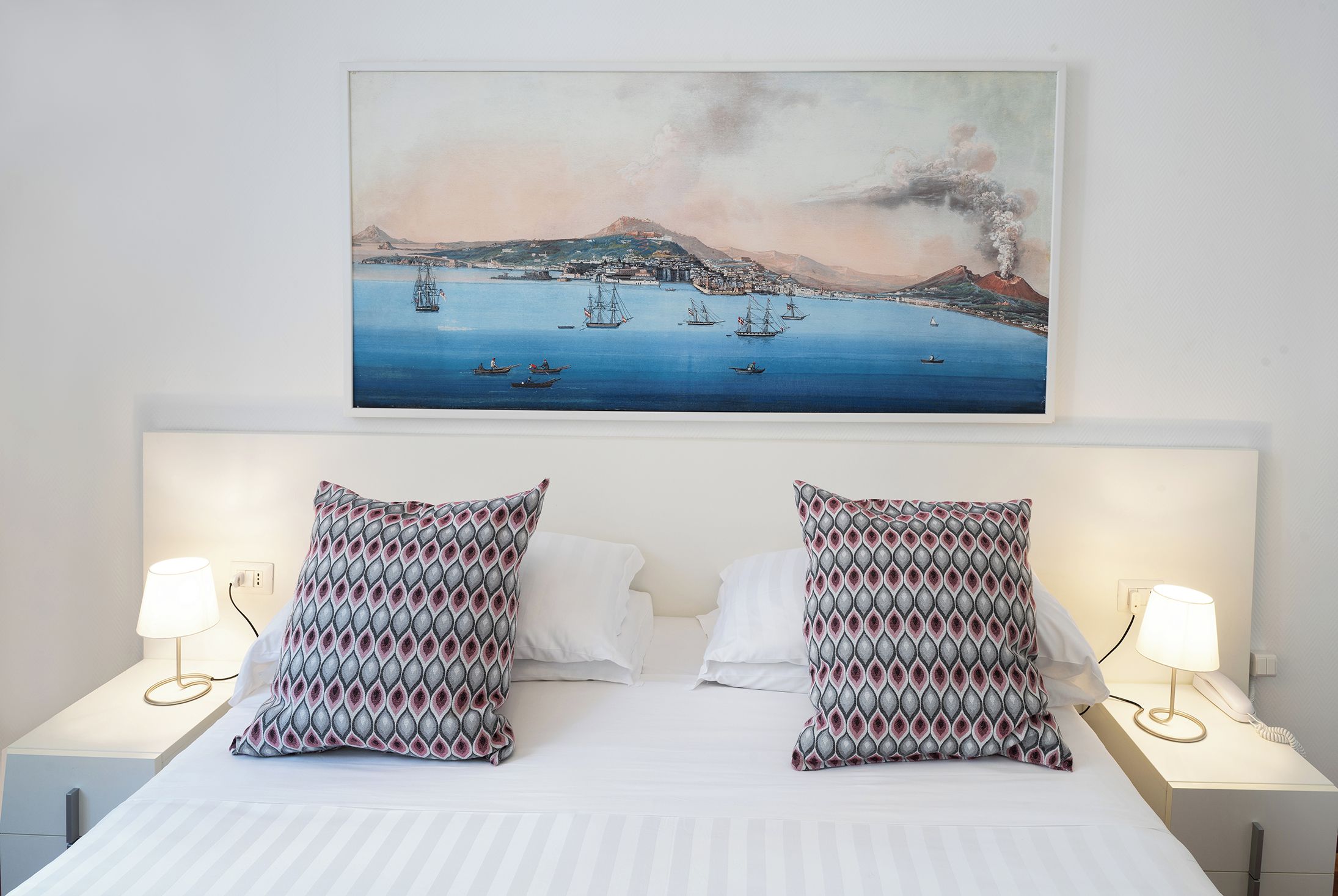 Ein Hotelzimmer mit einem großen Bett, zwei gemusterten Kissen, zwei Nachttischlampen und einem Bild einer Küstenlandschaft über dem Bett.