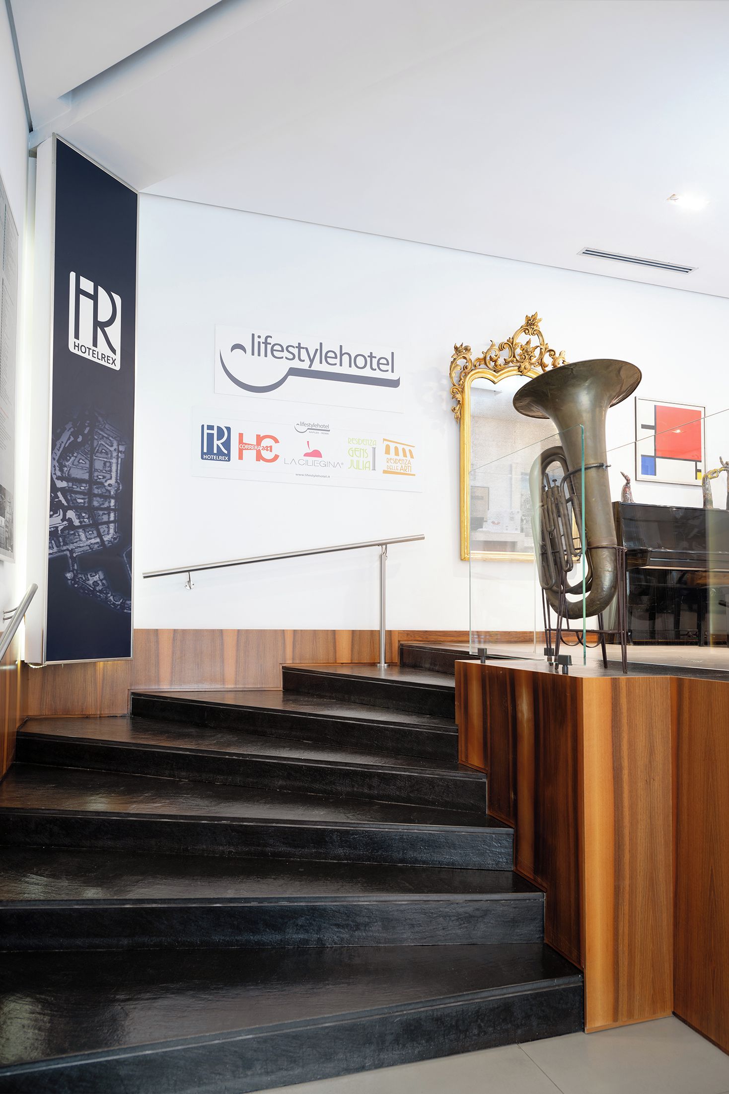 A reception desk with a brass instrument, a staircase, and a wall with various logos and a framed mirror.