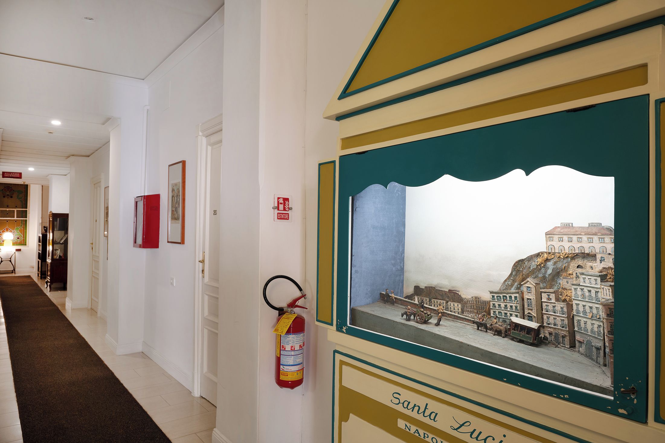Un couloir avec un extincteur fixé au mur, un tableau encadré représentant une scène historique et un panneau indiquant que l’on se trouve à « Santa Lucia ».