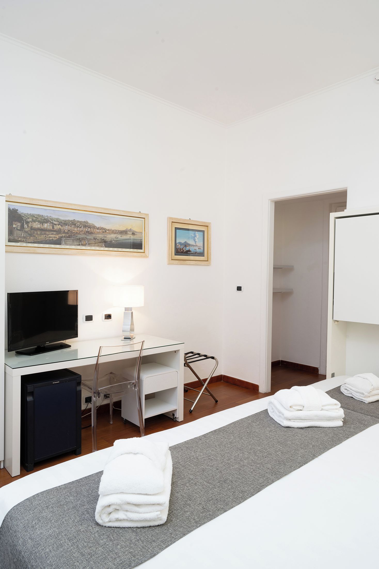 A hotel room with a bed, a desk, a chair, a TV, a lamp, and two framed pictures on the wall.