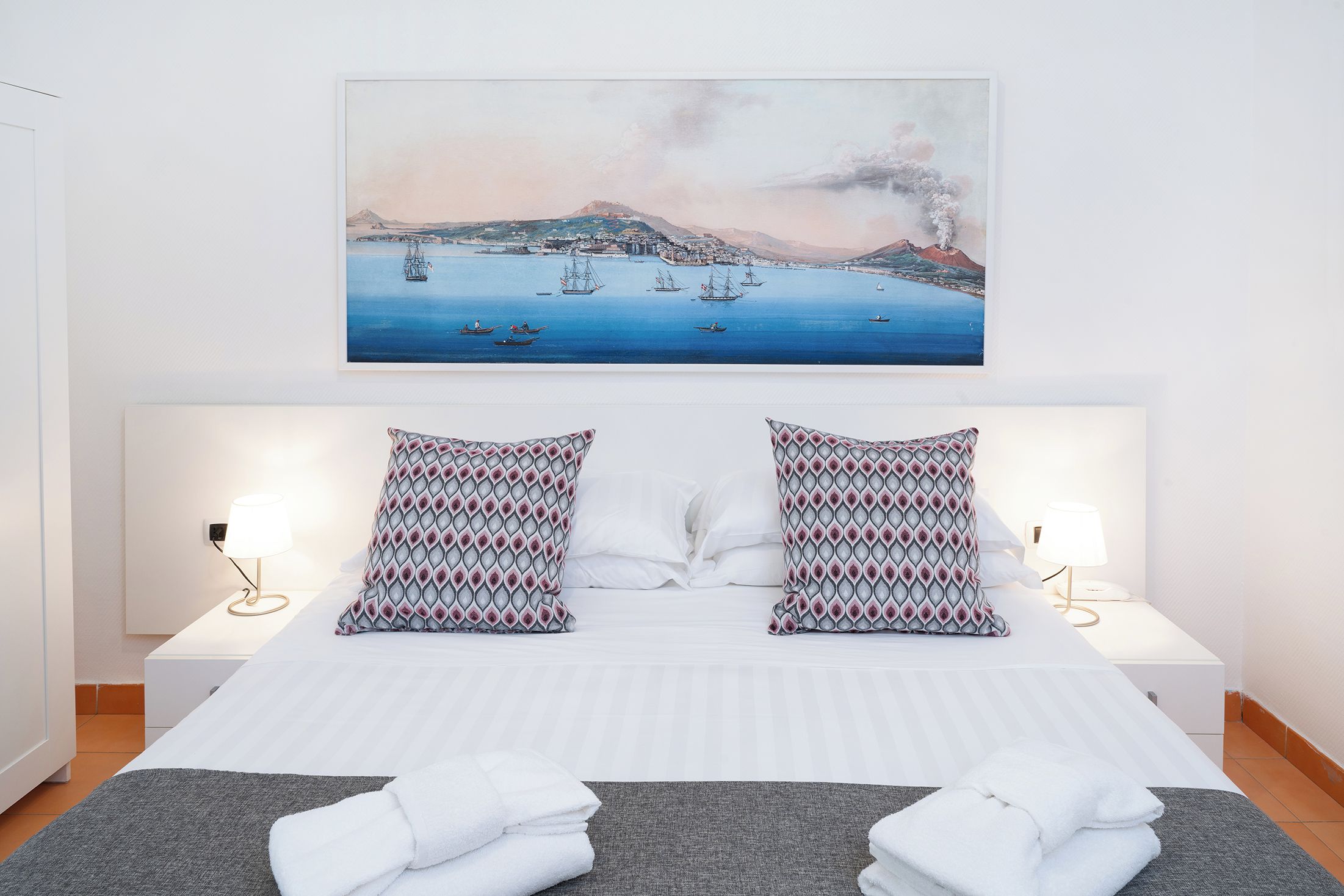 Ein Hotelzimmer mit einem Bett, zwei gemusterten Kissen, einem Gemälde mit einer Küstenansicht und Nachttischlampen.