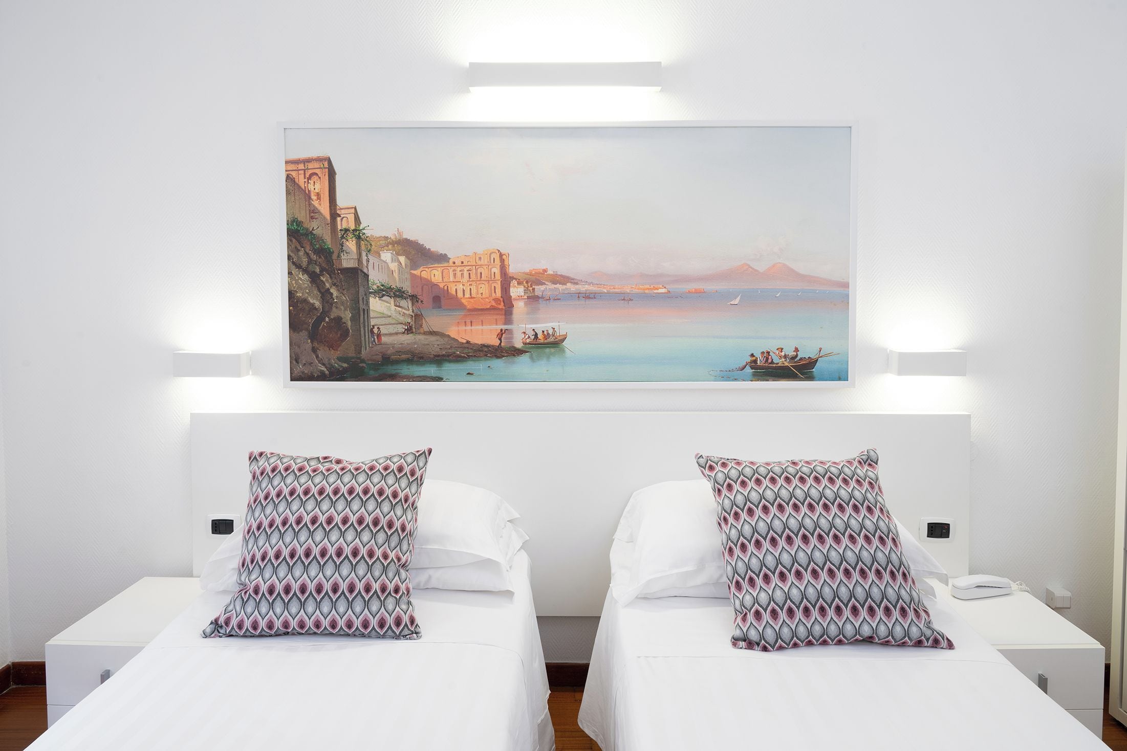 A hotel room with two beds, a large painting of a coastal scene, and two patterned pillows.