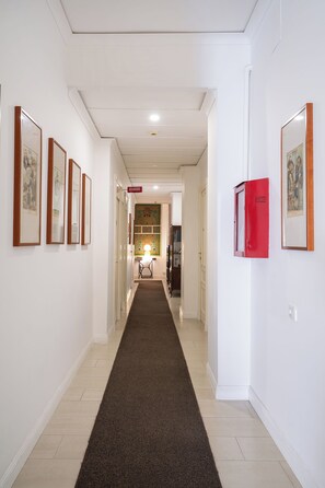 Een gang met kunstwerken in lijsten aan de wanden, een rode brandmeldknop en een vloerbedekking.