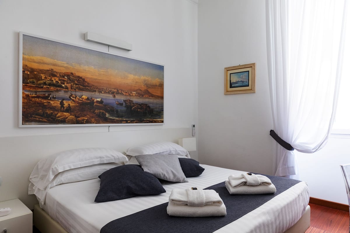 A hotel room with a bed, pillows, a painting, a small framed picture, and a window with curtains.