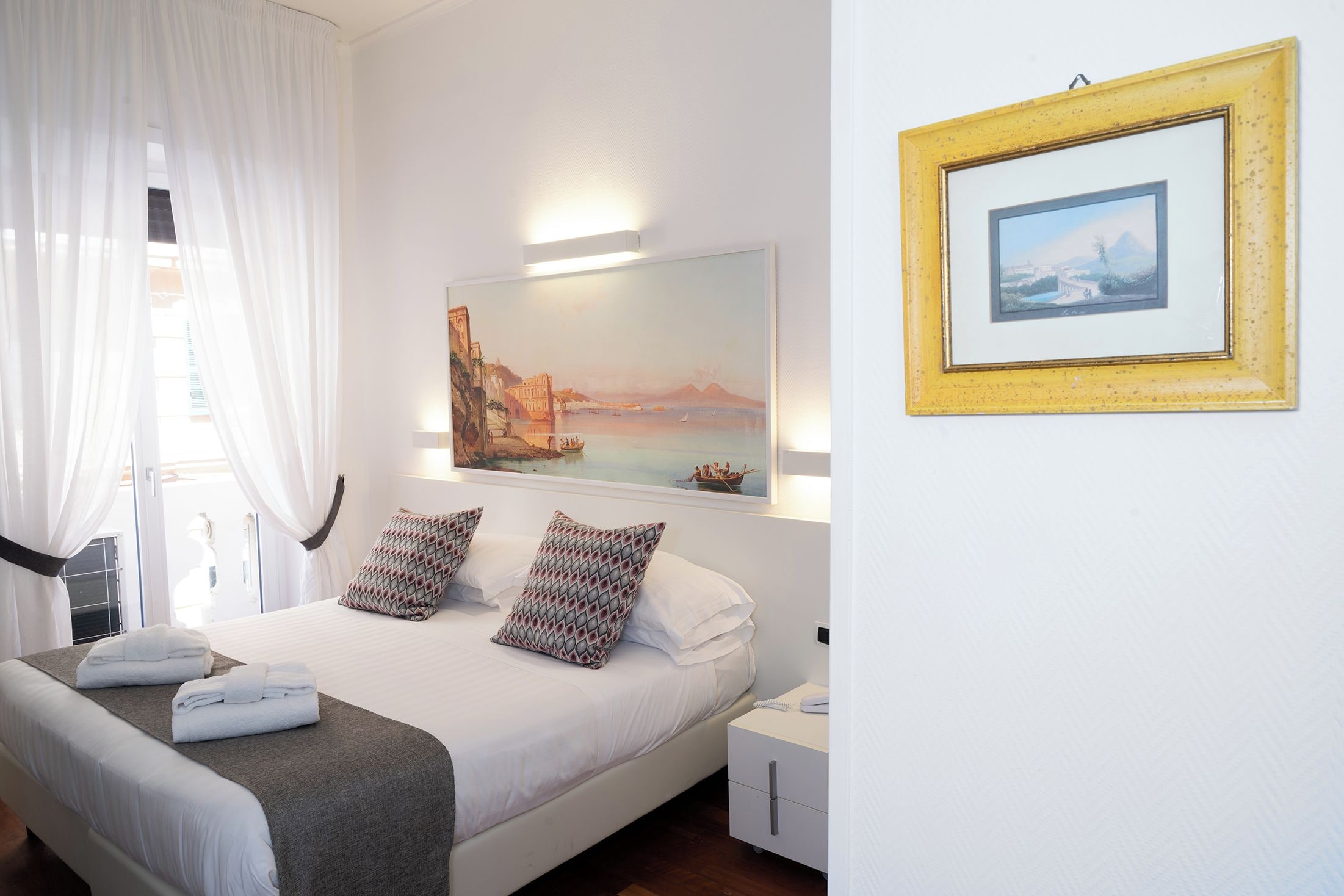 A hotel room with a bed, pillows, a bedside table, a nightstand, a framed picture, and a window with curtains.