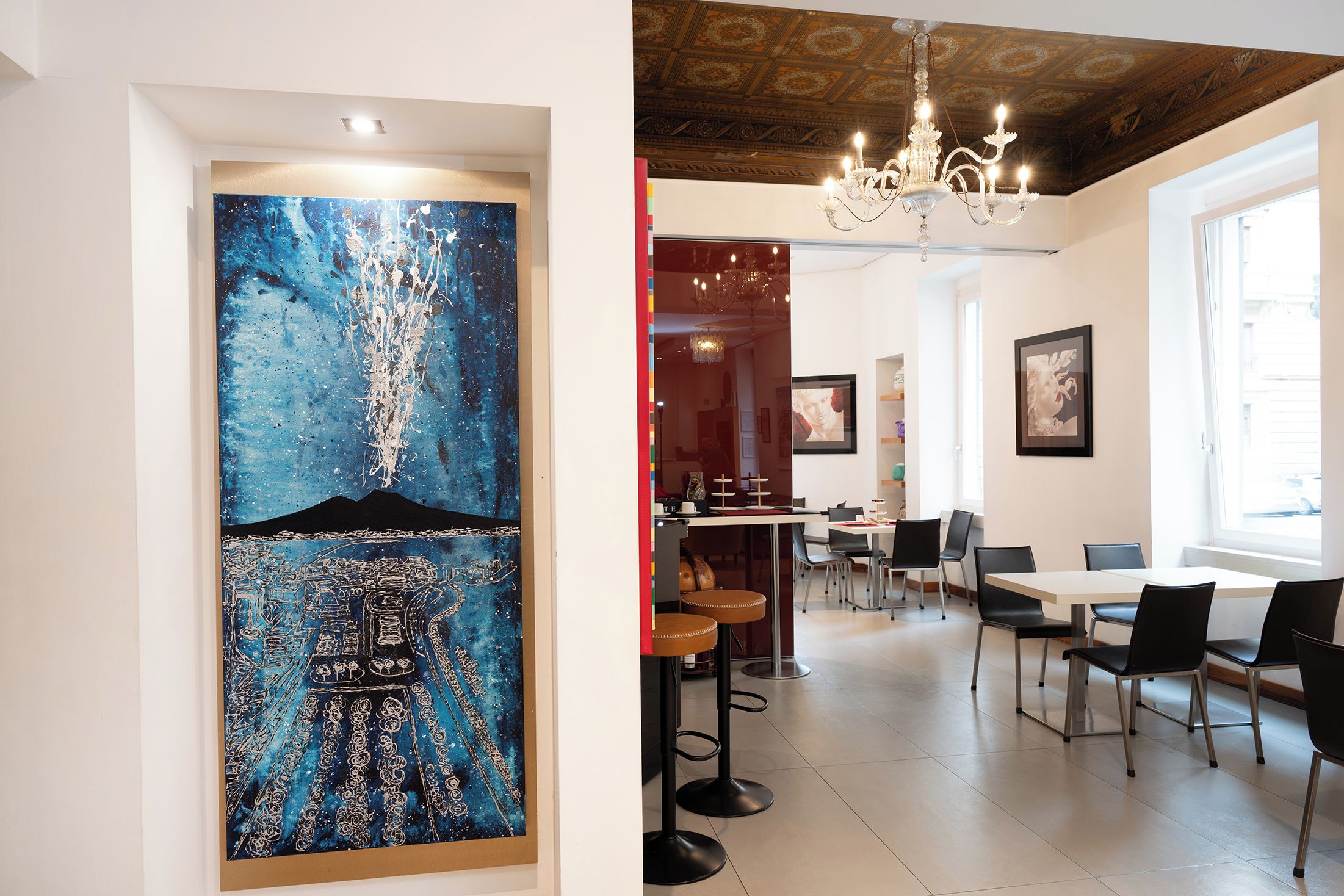 A dining area with a high ceiling, a chandelier, and a large abstract painting on the wall.