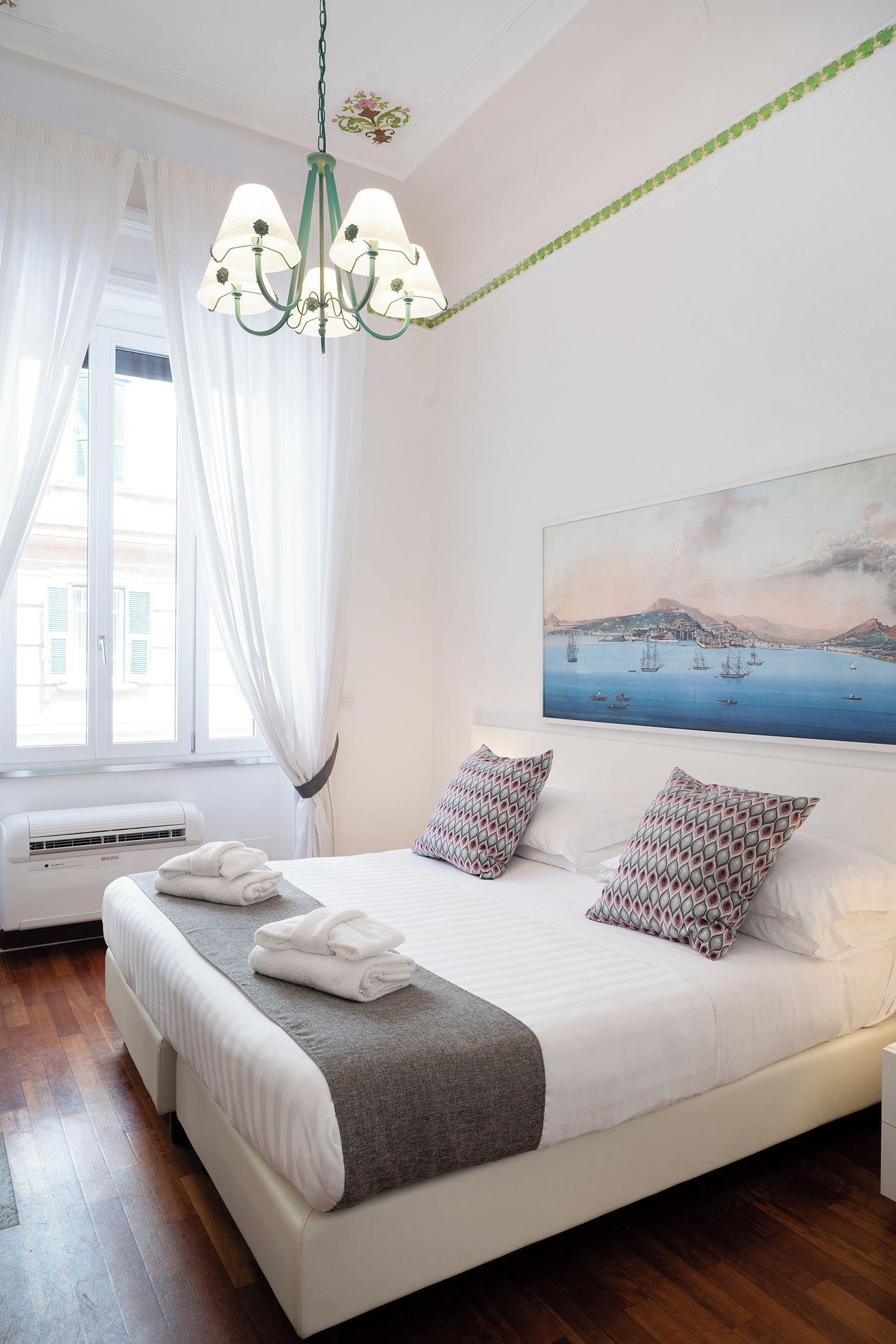 Een slaapkamer met een groot bed, een kroonluchter, een schilderij van een kustlandschap en een raam met gordijnen.