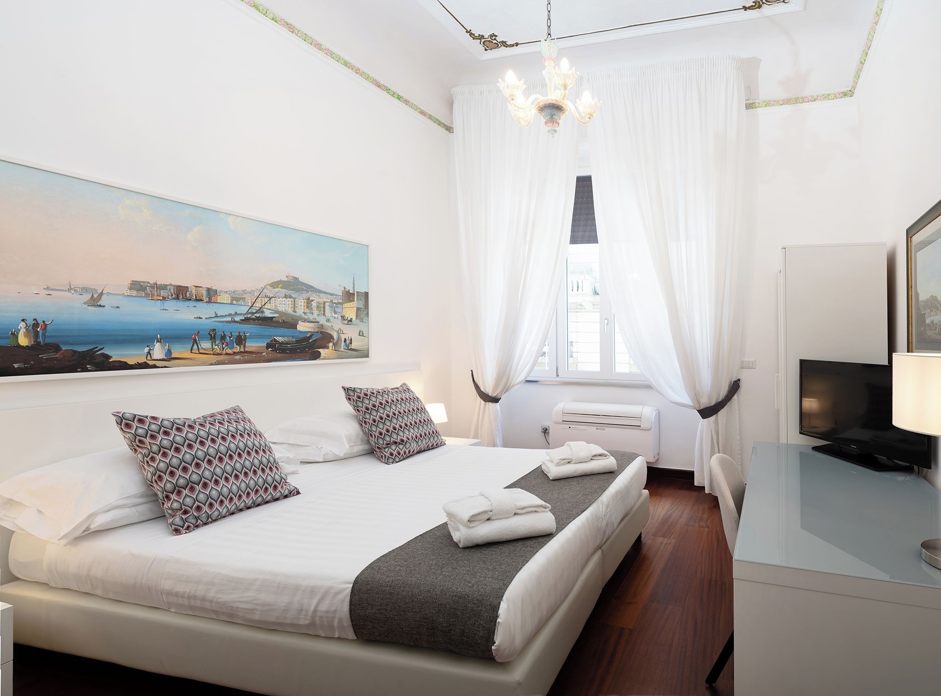 A hotel room with a bed, a TV, a painting, and a window with curtains.