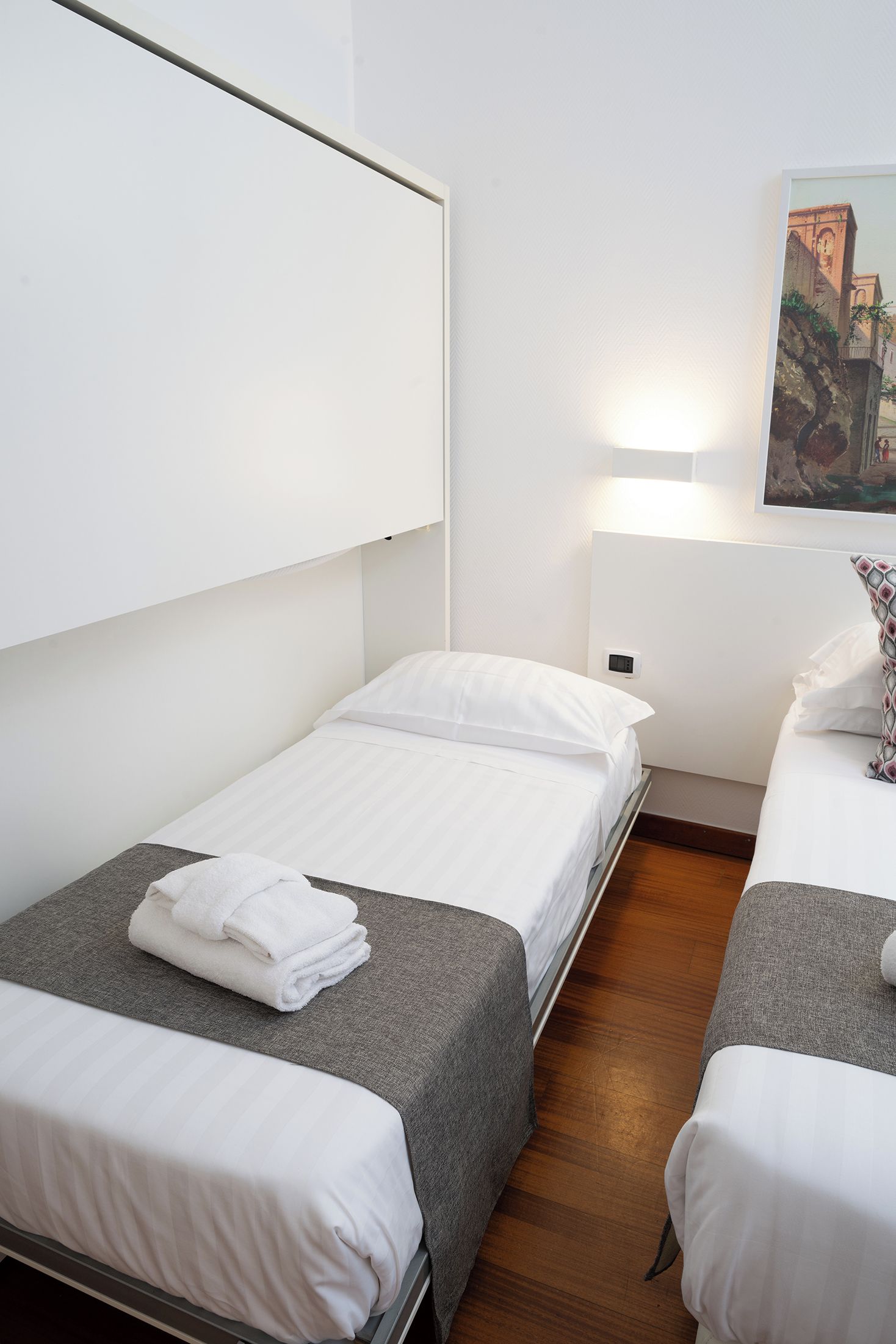 A hotel room with two single beds, a painting on the wall, and a bedside table with a lamp.