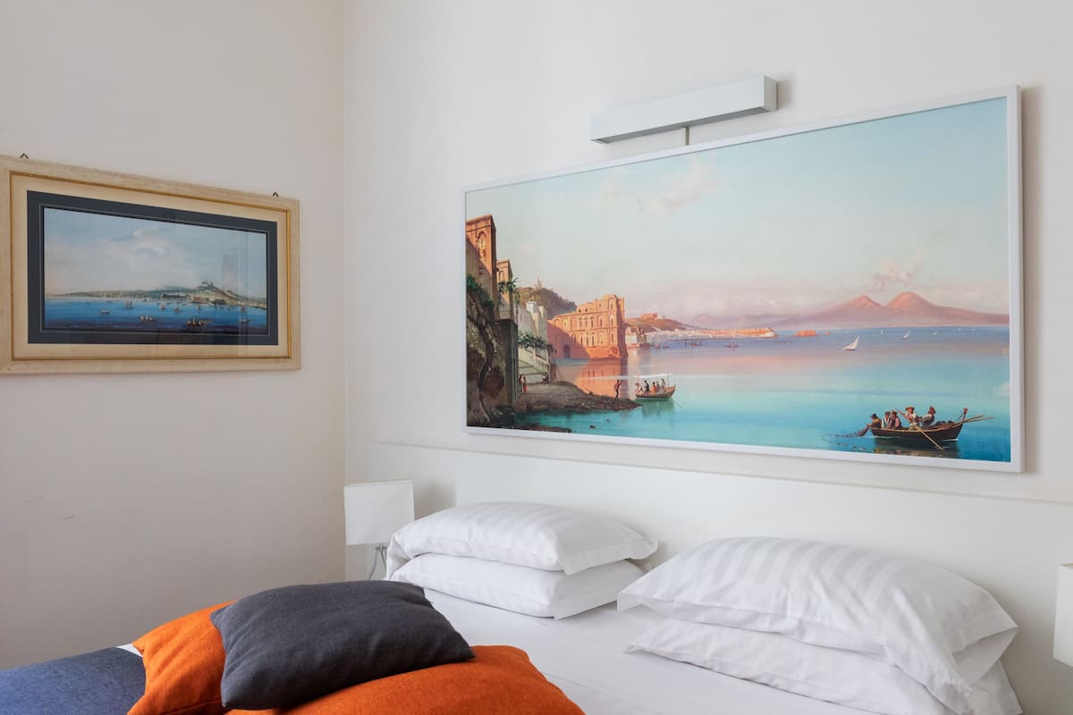 A hotel room with a large painting of a coastal scene, a bed with white and orange bedding, and a framed picture on the wall.
