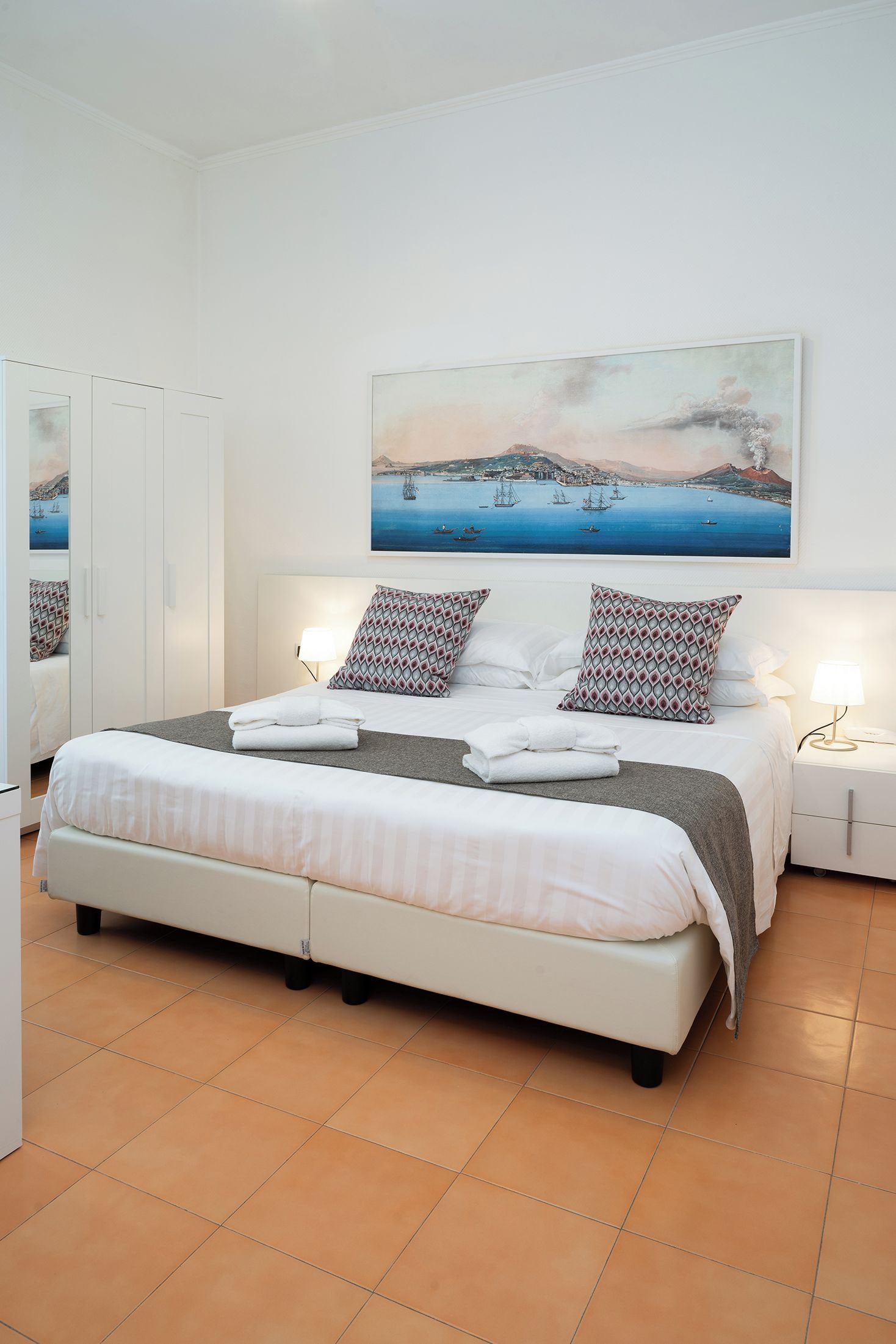 A bedroom with a large bed, two bedside tables, a nightstand, a painting of a coastal scene, and a wardrobe.
