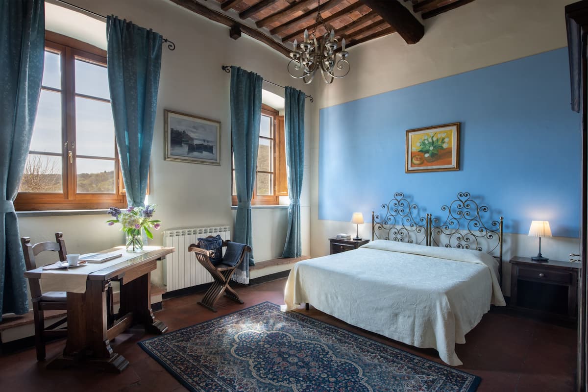 A bedroom with a bed, a desk with a chair, a window with curtains, a chandelier, and a painting on the wall.