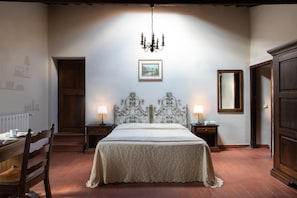 A bedroom with a large bed, two bedside tables with lamps, a chandelier, a wooden door, a framed picture on the wall, and a wooden wardrobe.