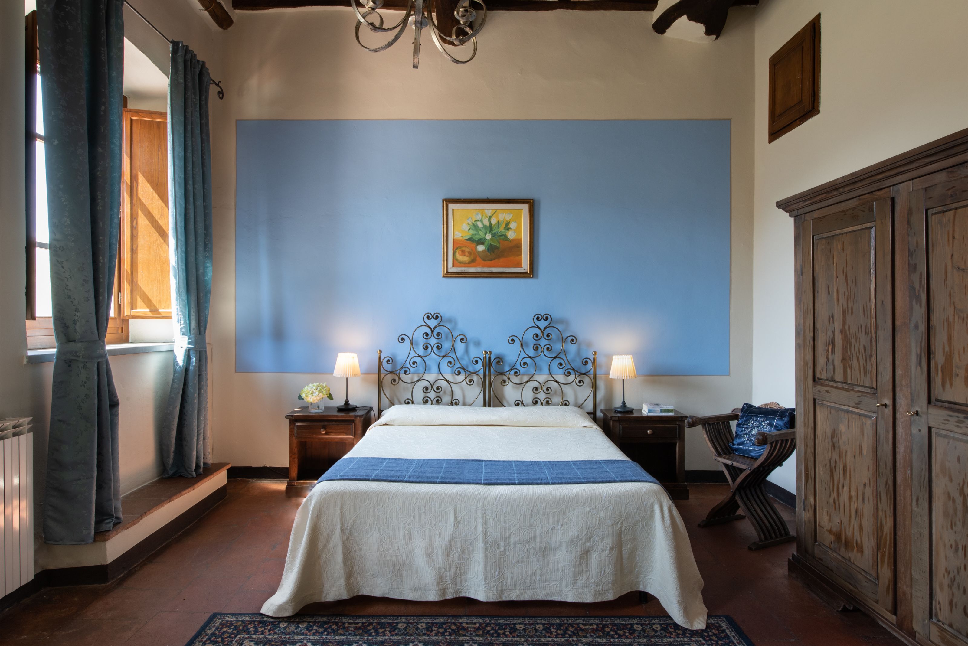 A bedroom with a bed, two bedside lamps, a blue wall, a framed picture, a wooden cabinet, and a window with curtains.