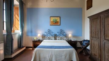 A bedroom with a bed, two bedside lamps, a blue wall, a framed picture, a wooden cabinet, and a window with curtains.