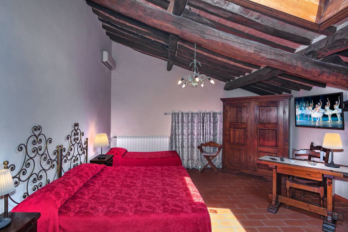 A bedroom with a red bedspread, a wooden desk with a lamp, a television, and a chandelier.