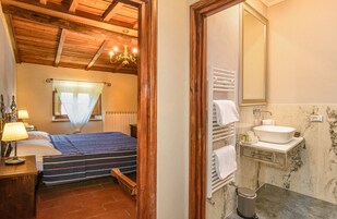 A bedroom with a bed, bedside table, lamp, and a window with a curtain. The room has wooden ceiling beams and a chandelier.