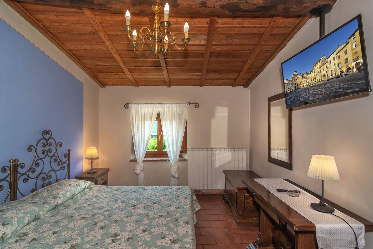 A bedroom with a wooden ceiling, a bed with a floral bedspread, a nightstand with a lamp, a desk with a lamp, a mirror, and a TV displaying a cityscape.