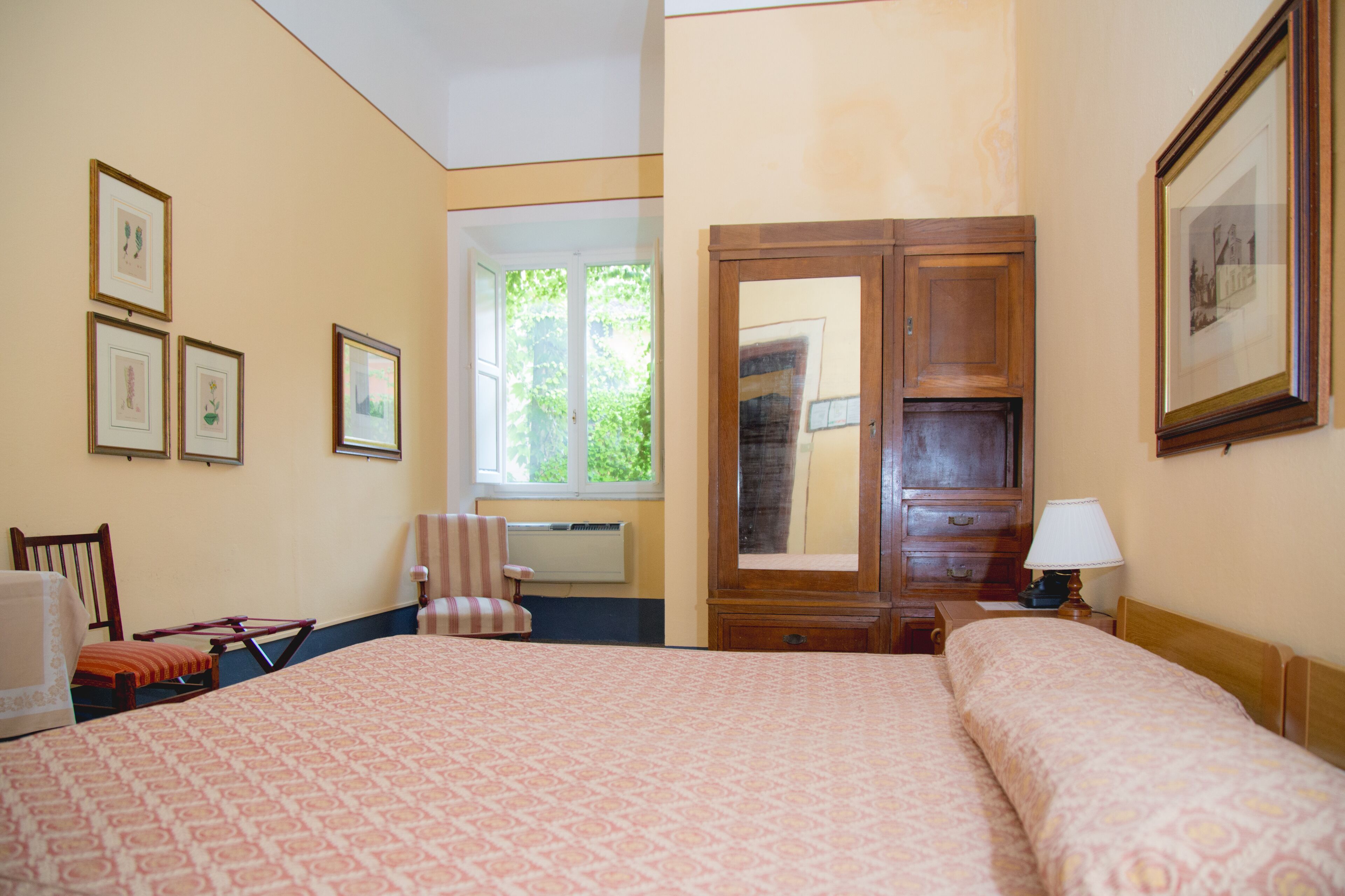 A hotel room with a large bed, a wooden wardrobe, a desk with a lamp, and a window with a view of greenery.