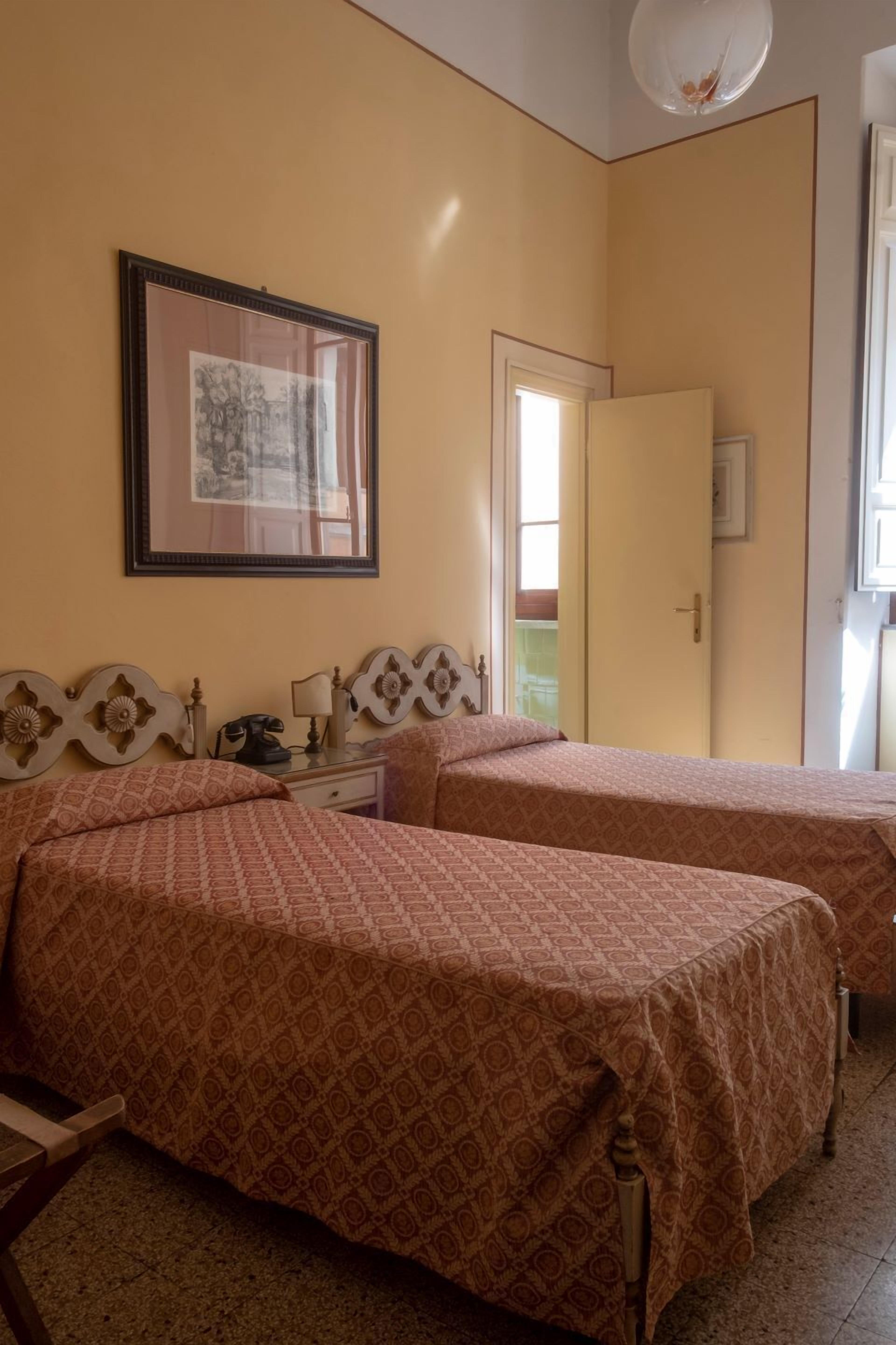 Une chambre avec deux lits, une table de chevet avec une lampe, un tableau encadré au mur et une porte donnant sur une autre pièce.