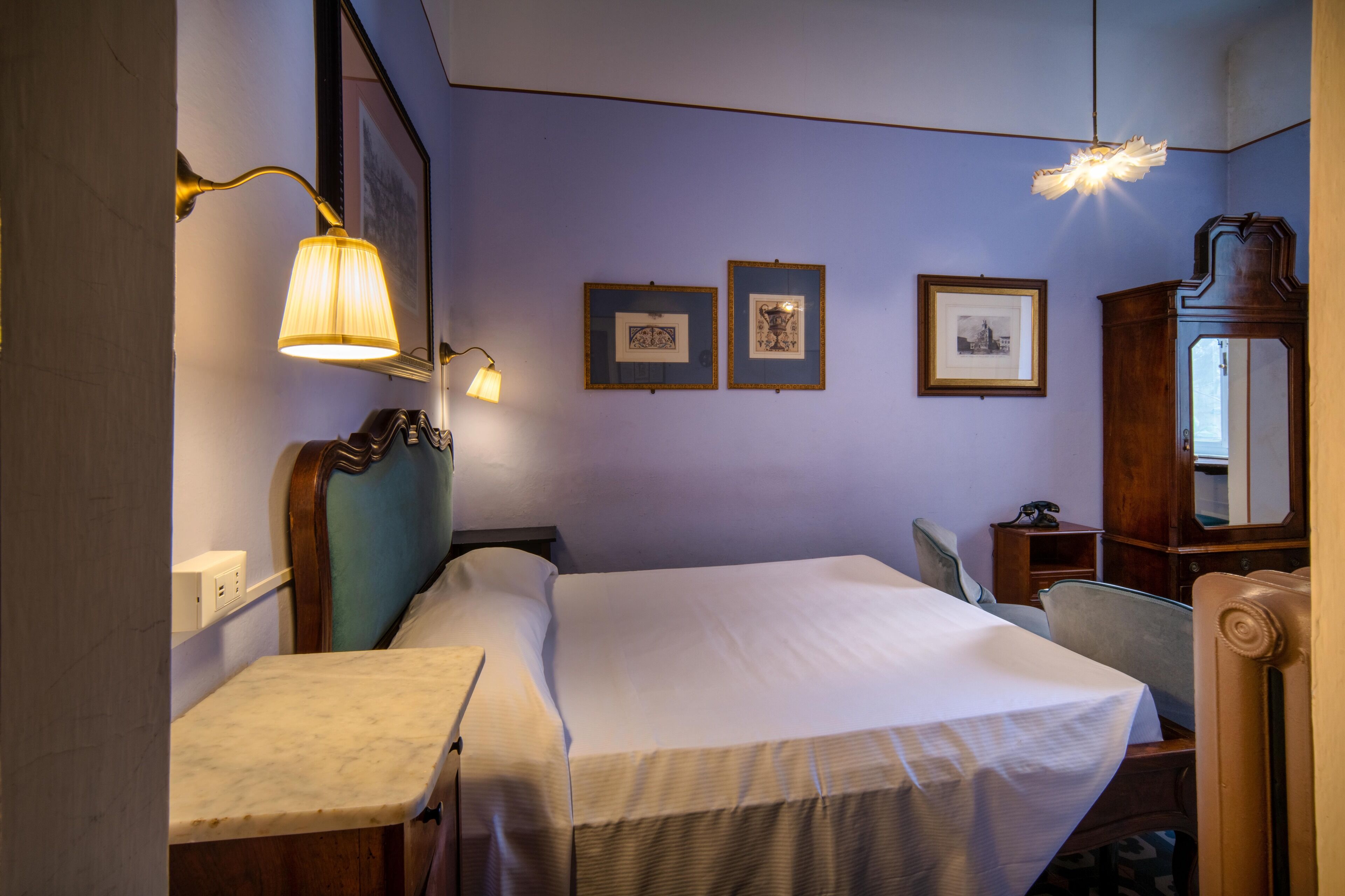 Una habitación de hotel con cama, mesita de noche, lámpara, cuadros en la pared y un armario de madera.
