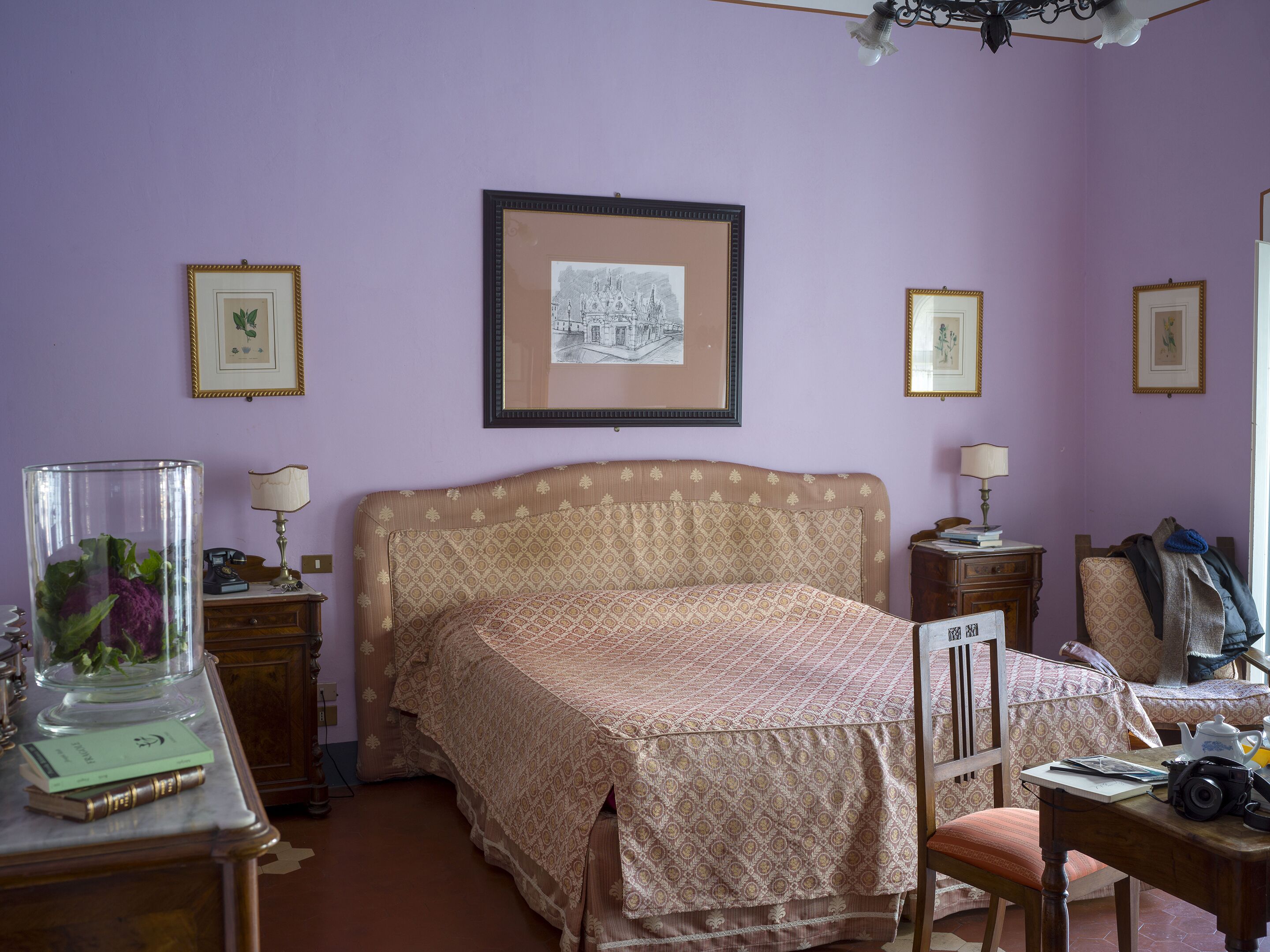 Un dormitorio con una colcha estampada, un cuadro en la pared, una mesita de noche con una lámpara y una silla con una manta.