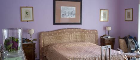 Un dormitorio con una colcha estampada, un cuadro en la pared, una mesita de noche con una lámpara y una silla con una manta.