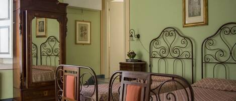 Un dormitorio con piso a cuadros, una cama con dosel de madera, dos camas de metal con cabeceros ornamentados, una silla de madera y una mesita de noche con lámpara.
