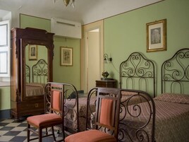 Un dormitorio con piso a cuadros, una cama con dosel de madera, dos camas de metal con cabeceros ornamentados, una silla de madera y una mesita de noche con lámpara.
