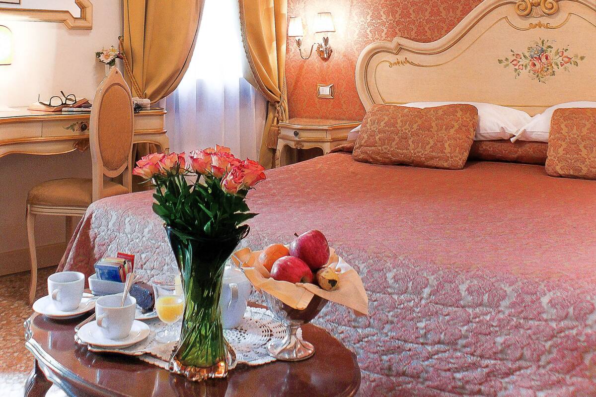 A hotel room with a bed, a desk, a chair, a vase with flowers, and a fruit basket.
