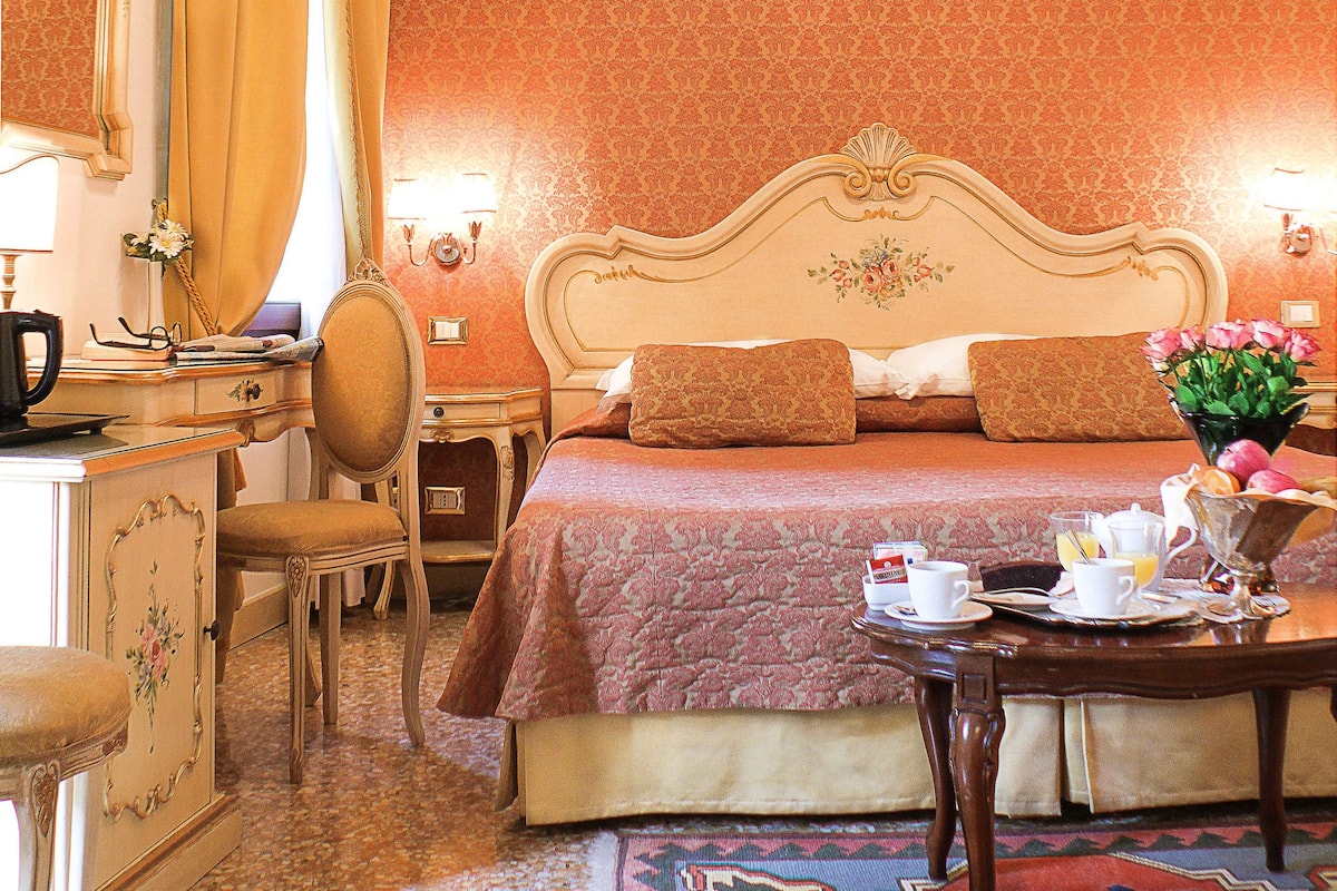 A hotel room with a wooden ceiling, a large bed with a floral headboard, a desk with a mirror, and a small table with a vase of flowers.