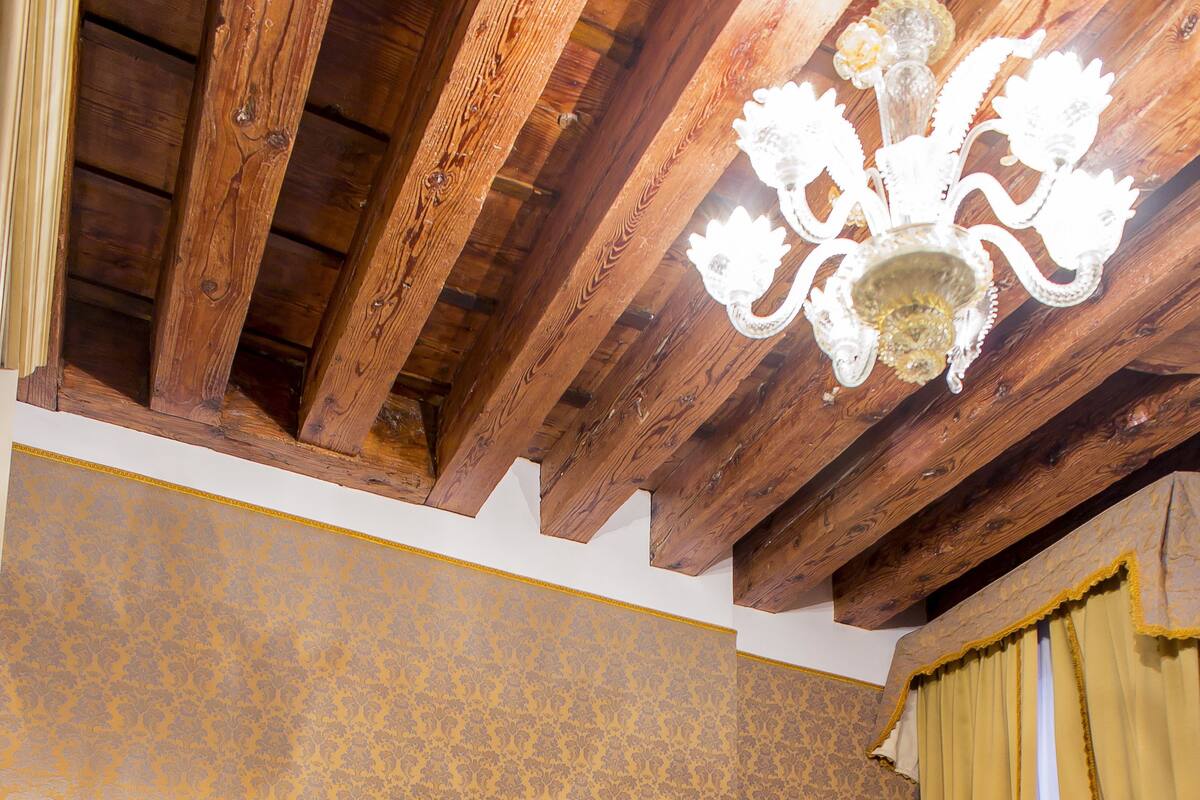 A hotel room with a wooden ceiling, a bed with a floral headboard, a nightstand with a lamp, and a curtain.