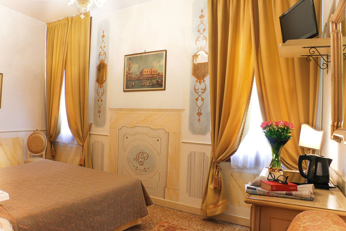 A hotel room with a bed, a desk with a lamp, a TV, a vase with flowers, and a painting on the wall.