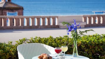 A table with pastries, drinks, and a vase of flowers, set against a backdrop of a beach and ocean view.