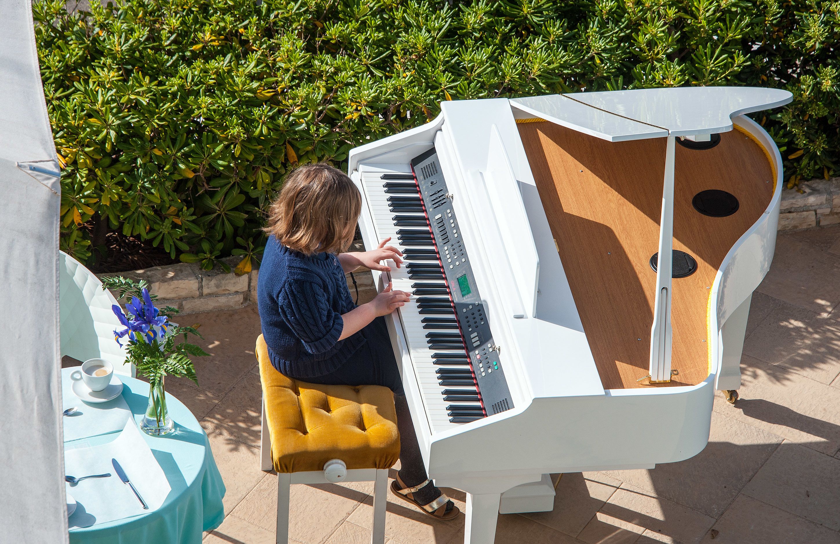 En person spelar på ett vitt piano utomhus, med ett bord och blommor i närheten.