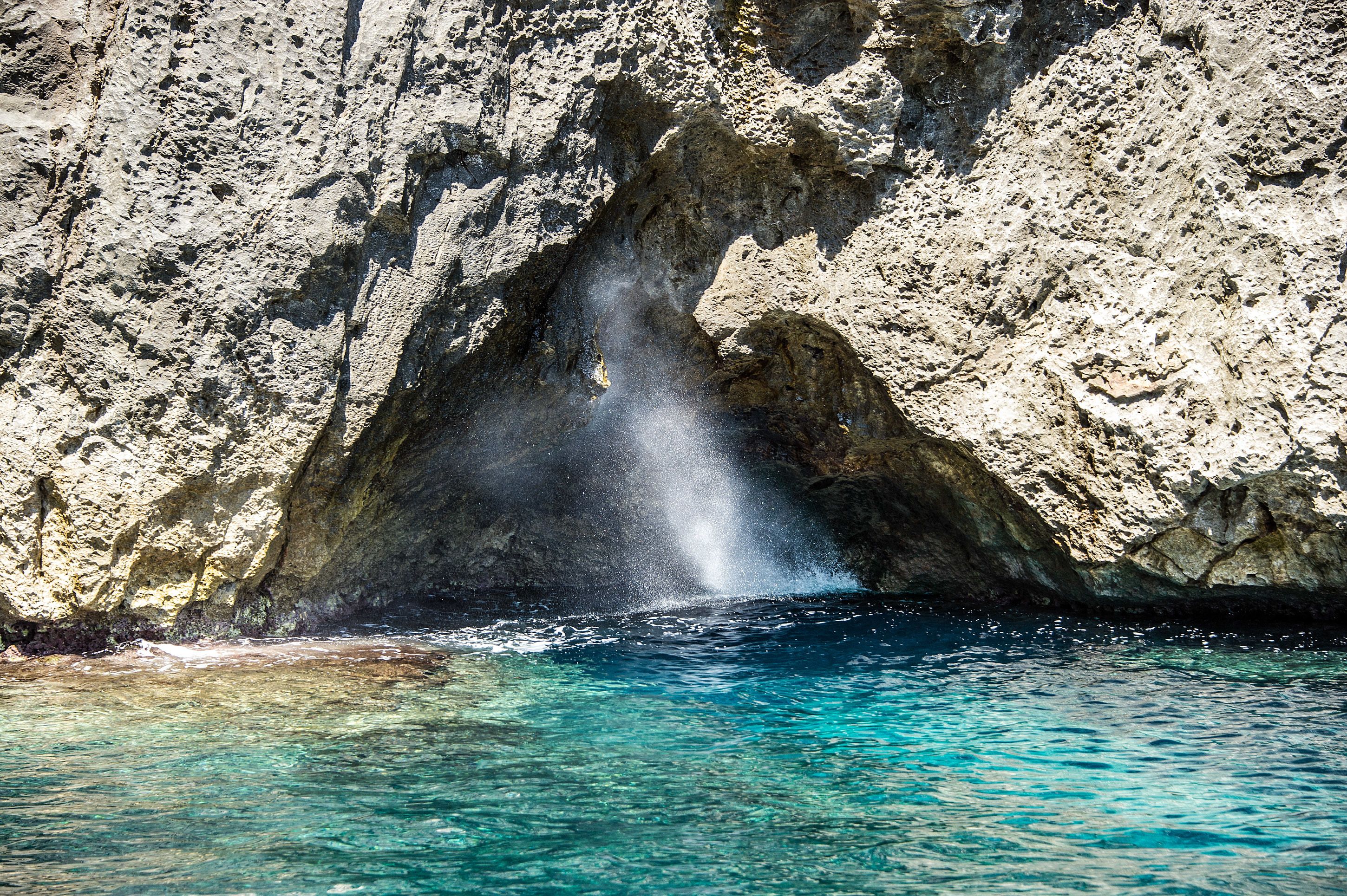 Escena costera rocosa con agua azul cristalina y una visible columna de agua.