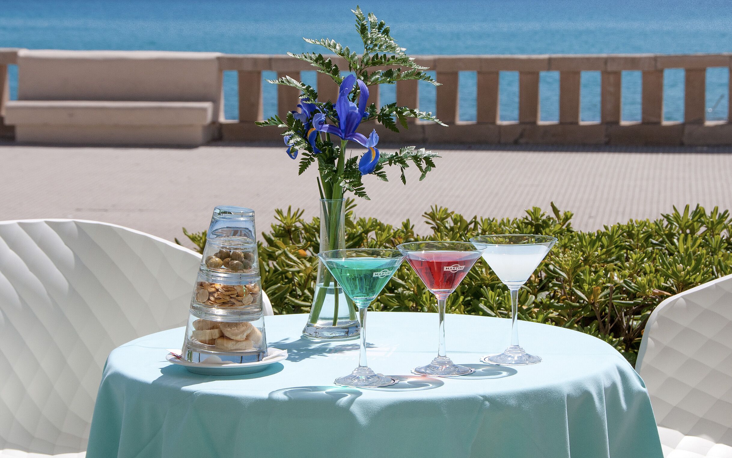 Una mesa con un florero de vidrio transparente, una copa de martini y un vaso de postre sobre un mantel, con vista al mar de fondo.