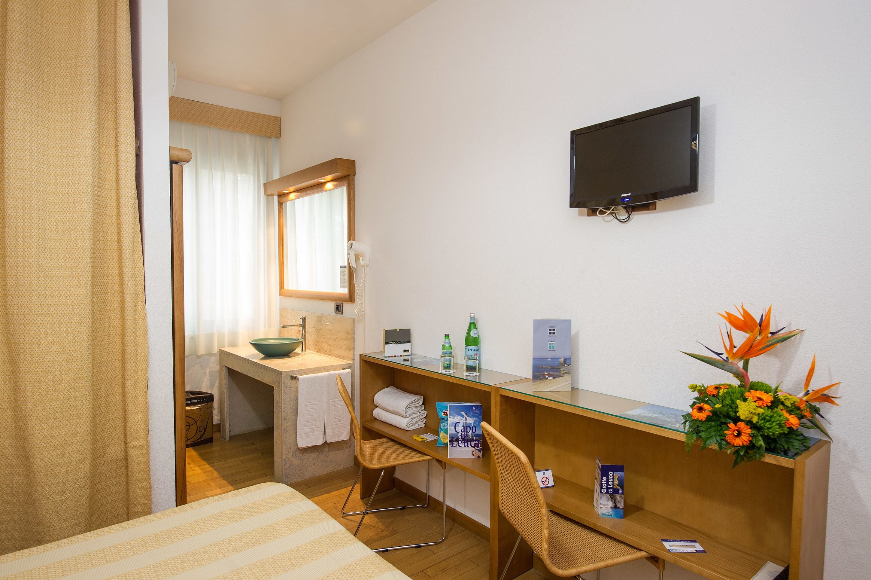 A hotel room with a bed, a desk with a chair, a television, a mirror, a sink, and a window with curtains.