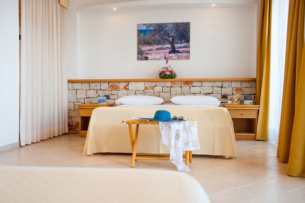 A neatly arranged bedroom with a bed, bedside tables, a small table with a hat, and a painting on the wall.