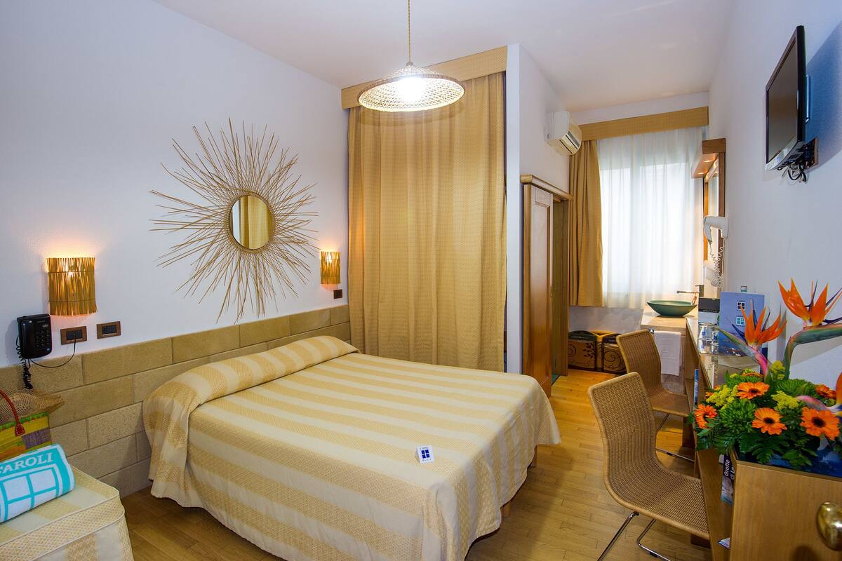 A hotel room with a bed, a desk, a chair, a mirror, and a wall-mounted TV.