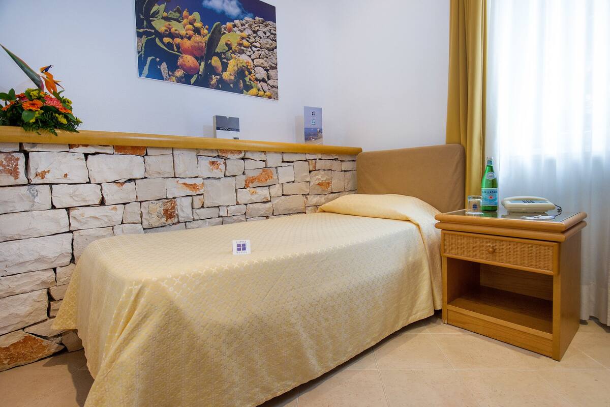 A single bed with a beige bedspread, a wooden nightstand with a lamp, a bottle of water, and a framed picture on the wall.