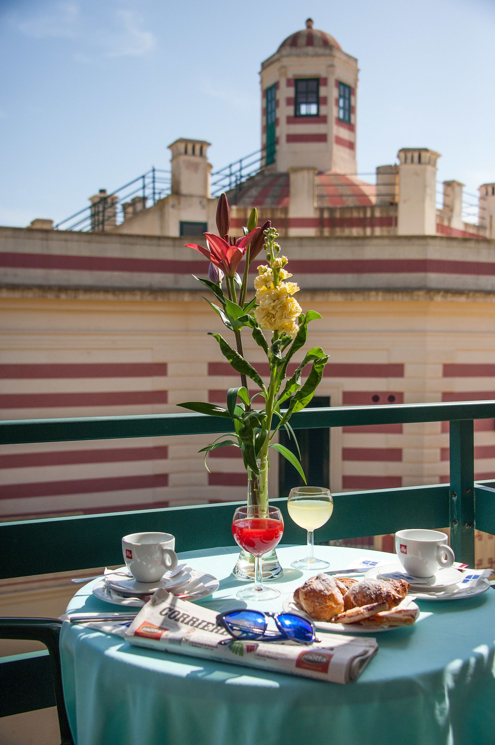Une table avec un journal, une tasse, un verre de vin rouge, un verre de vin blanc et des pâtisseries, le tout placé sur un balcon d’où l’on peut admirer un immeuble aux murs rayés et surmonté d’une tour.