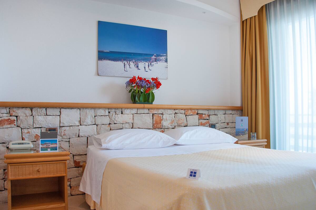 A hotel room with a stone wall, a bed with white linens, a nightstand with a phone, a painting of a beach scene, and a vase with flowers.