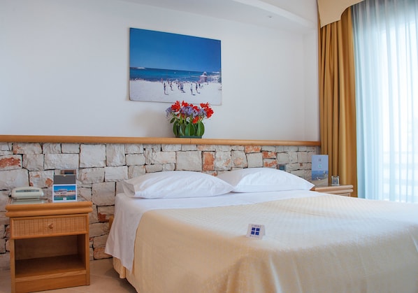 A hotel room with a stone wall, a bed with white linens, a nightstand with a phone, a painting of a beach scene, and a vase with flowers. - Hotel Terminal - Caroli Hotels (Castrignano del Capo)