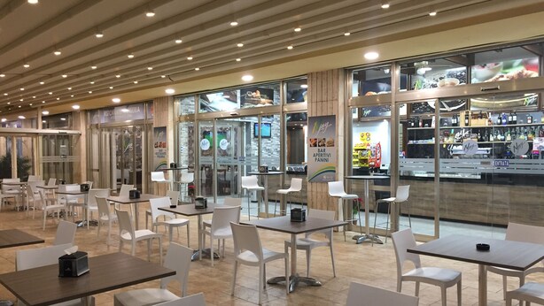 Un interior de restaurante moderno con sillas y mesas blancas, ventanas amplias y un área de bar visible a través del vidrio.
