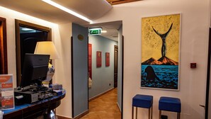A hotel lobby with a reception desk, a computer, a lamp, a painting of a whale, and two blue bar stools. - Hotel Neapolis (Naples)