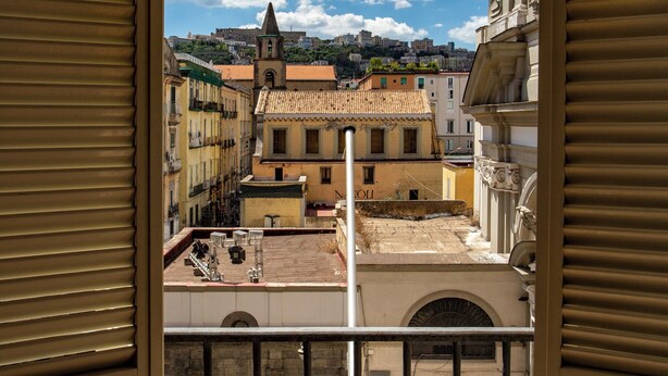 A view from a window overlooking a historic town square with buildings, a church, and a hill in the background. - Hotel Neapolis (Naples)