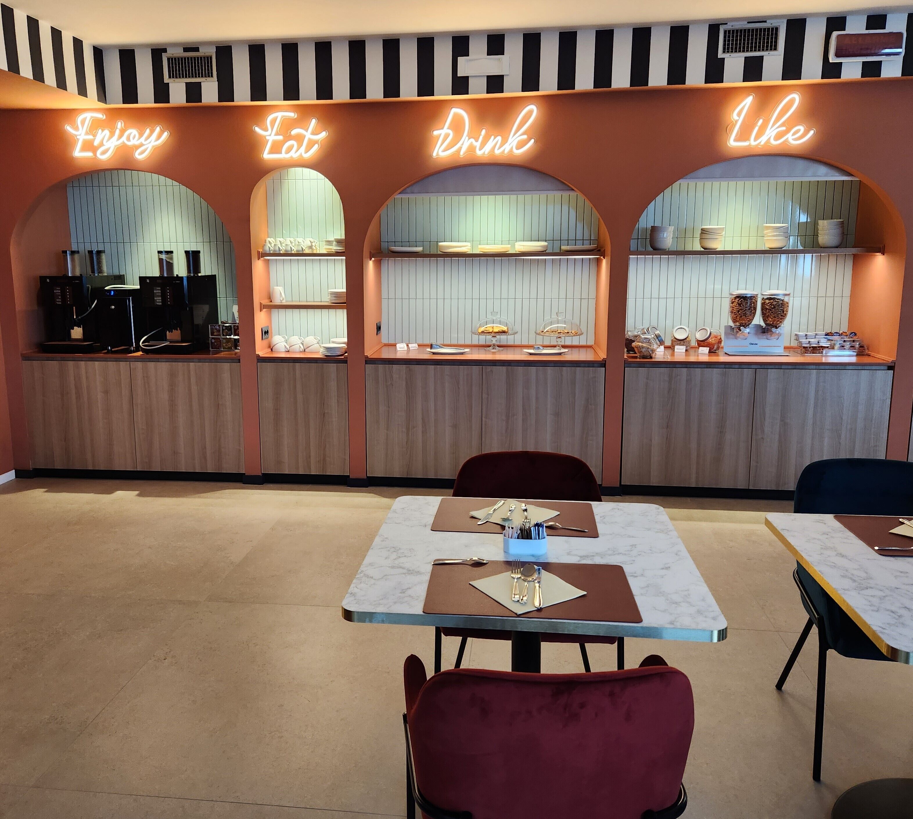 A modern cafe with neon signs reading "Enjoy," "Eat," "Drink," and "Like." There are espresso machines, a variety of food and drink dispensers, and neatly arranged tables with chairs.